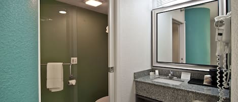 Une salle de bain avec une douche à parois de verre, des toilettes blanches, un lavabo sur un plan de travail en granit, un sèche-cheveux et un miroir.