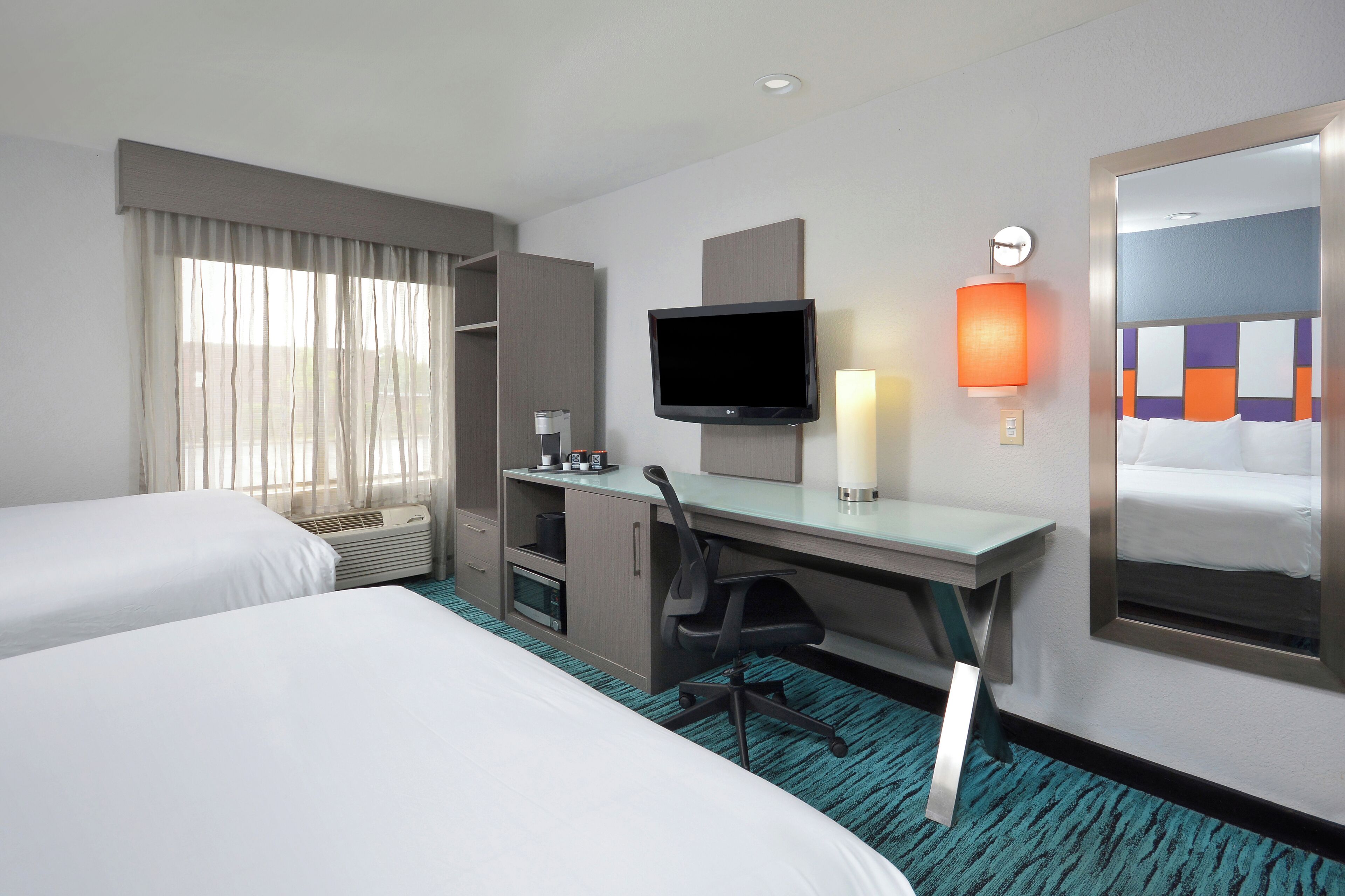 Une chambre d’hôtel avec deux lits, un bureau avec une chaise, une télévision, une lampe et un grand miroir.