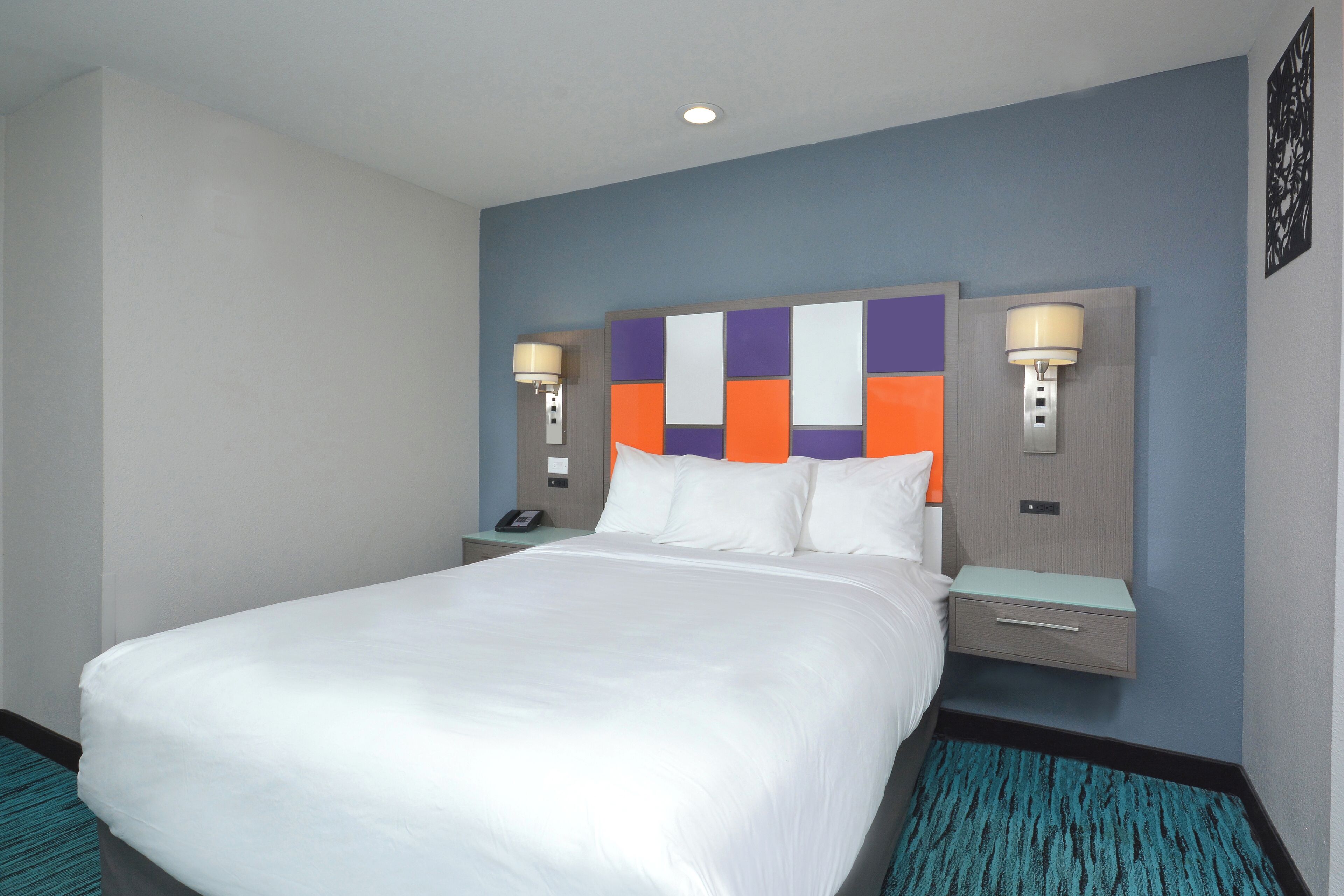 Quarto de hotel com uma cama grande, duas mesas de cabeceira com abajures, um criado-mudo e um tapete azul.