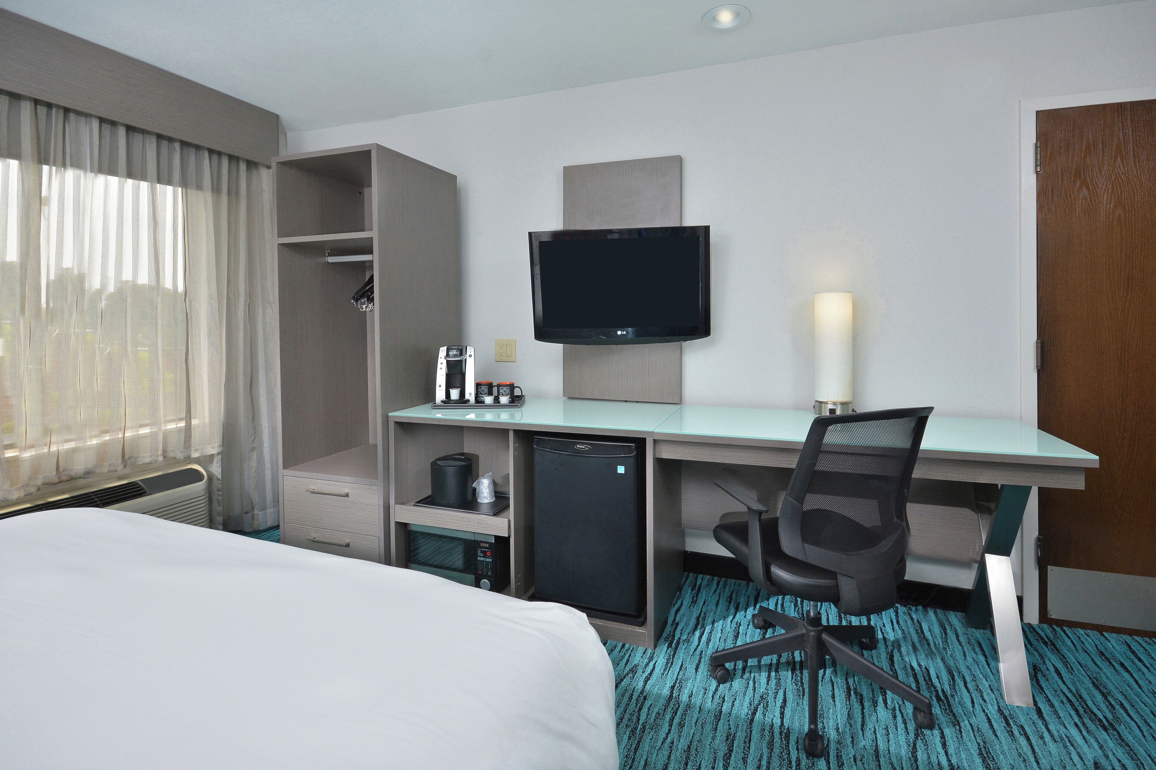 A hotel room with a bed, a desk with a computer, a TV, a mini-fridge, a coffee maker, and a lamp.