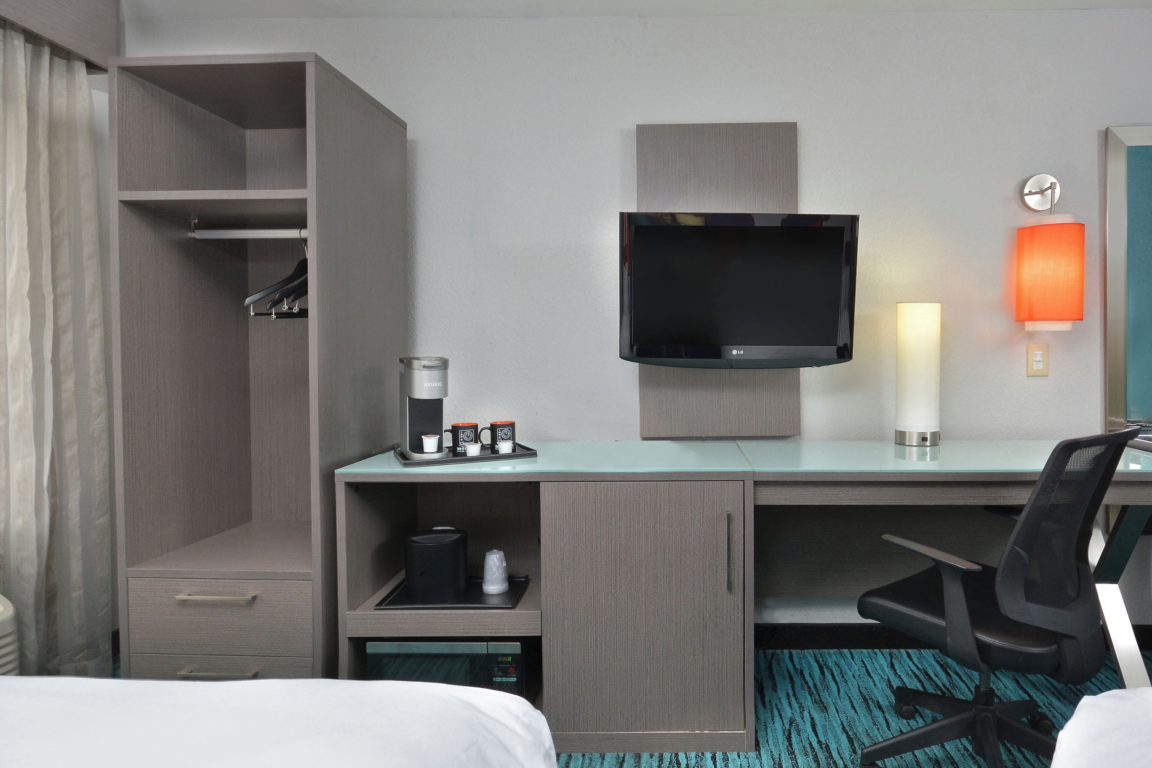 Una habitación de hotel con cama, escritorio, televisor, cafetera y lámpara.
