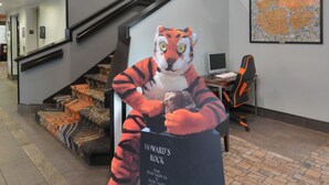 Une mascotte en forme de tigre tenant une pancarte sur laquelle on peut lire « HOWARD’S ROCK », un escalier et un bureau avec un ordinateur.