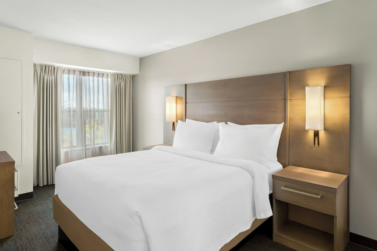 A hotel room with a large bed, two bedside lamps, a wooden nightstand, and a window with curtains.