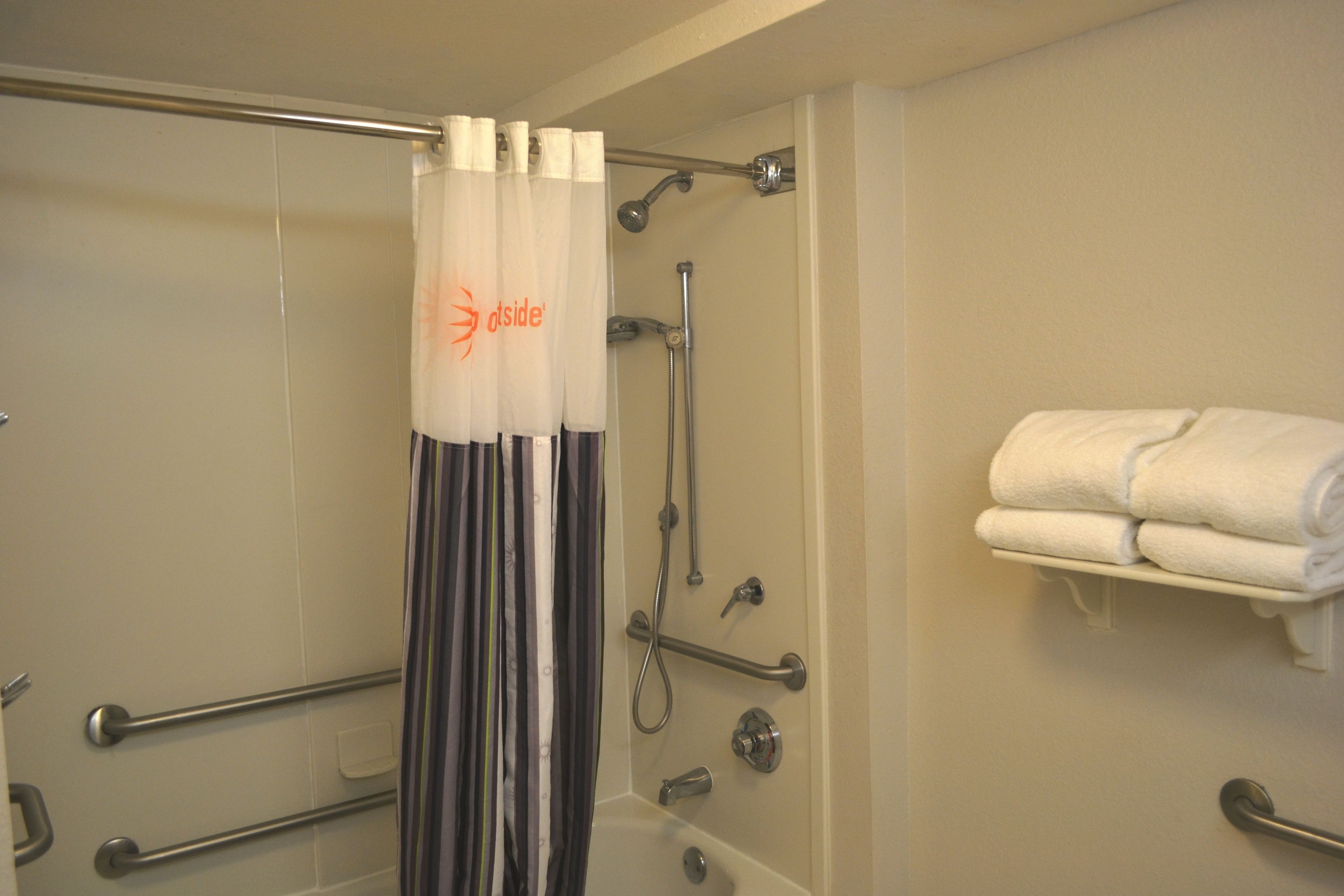 A bathroom with a shower, grab bars, a towel rack, and a curtain with a logo.