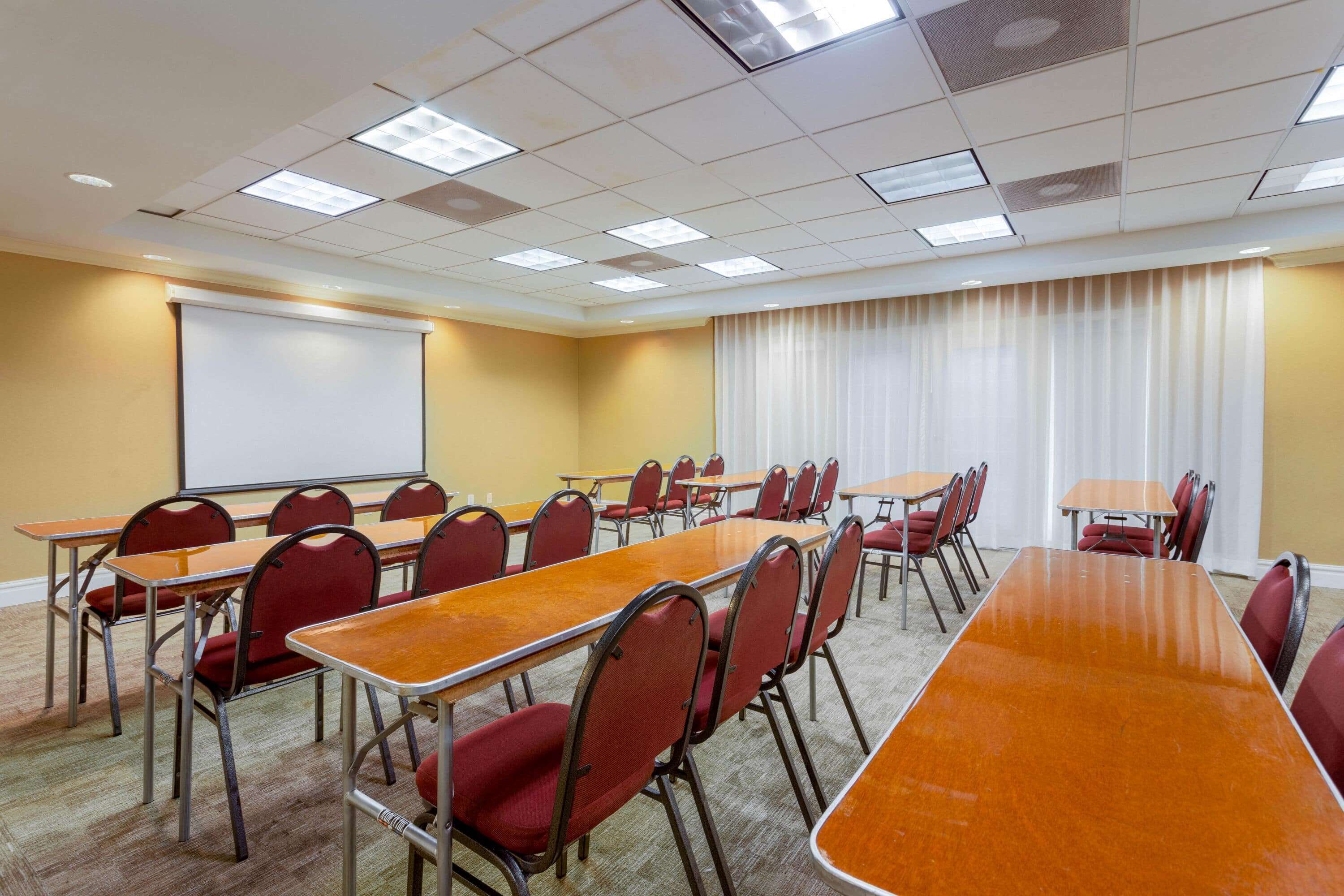 Un salón de conferencias con varias mesas y sillas, una pantalla de proyector y un ventanal con cortinas.