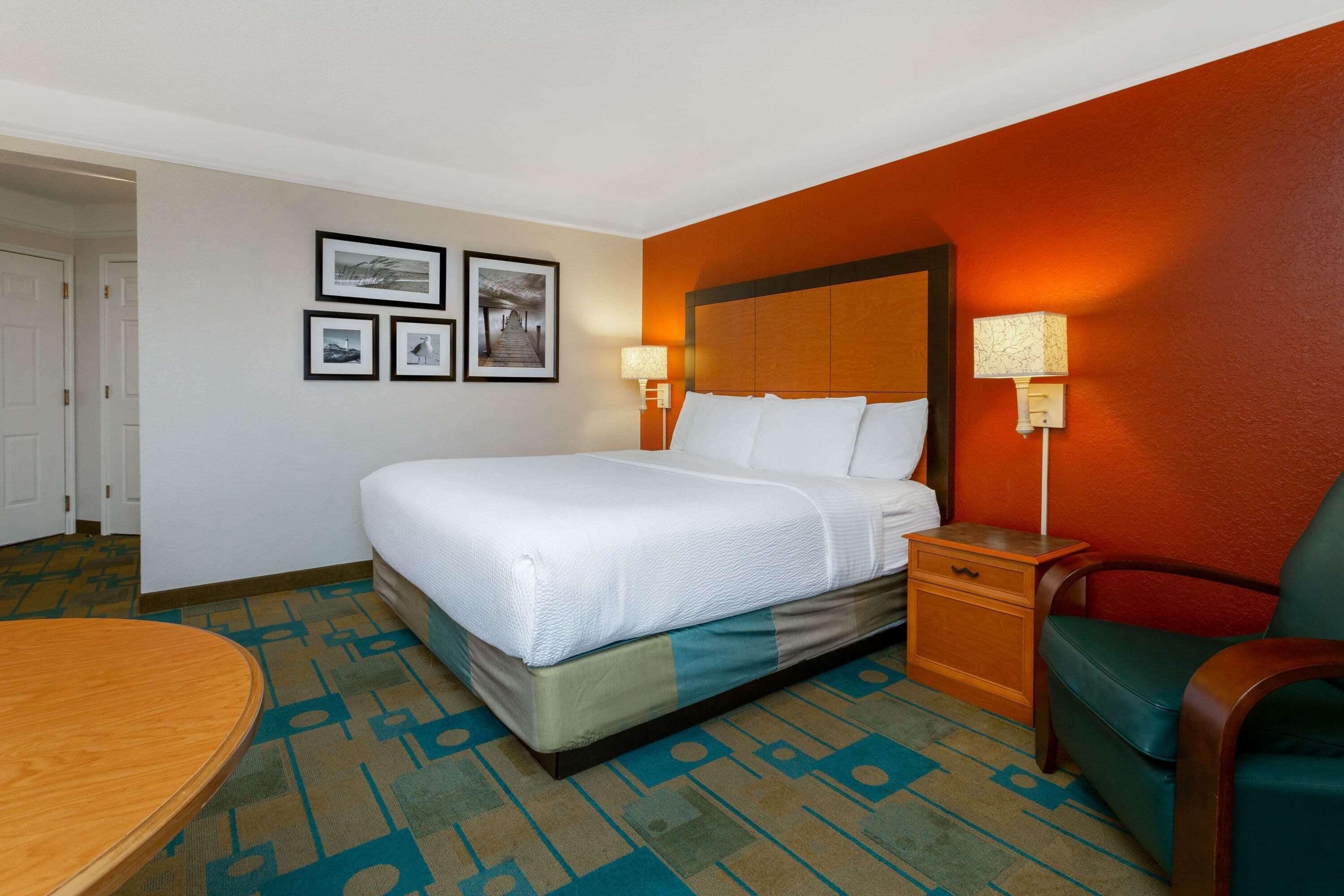 A hotel room with a bed, a nightstand, a chair, and a table. There are framed pictures on the wall.