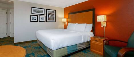 A hotel room with a bed, a nightstand, a chair, and a table. There are framed pictures on the wall.