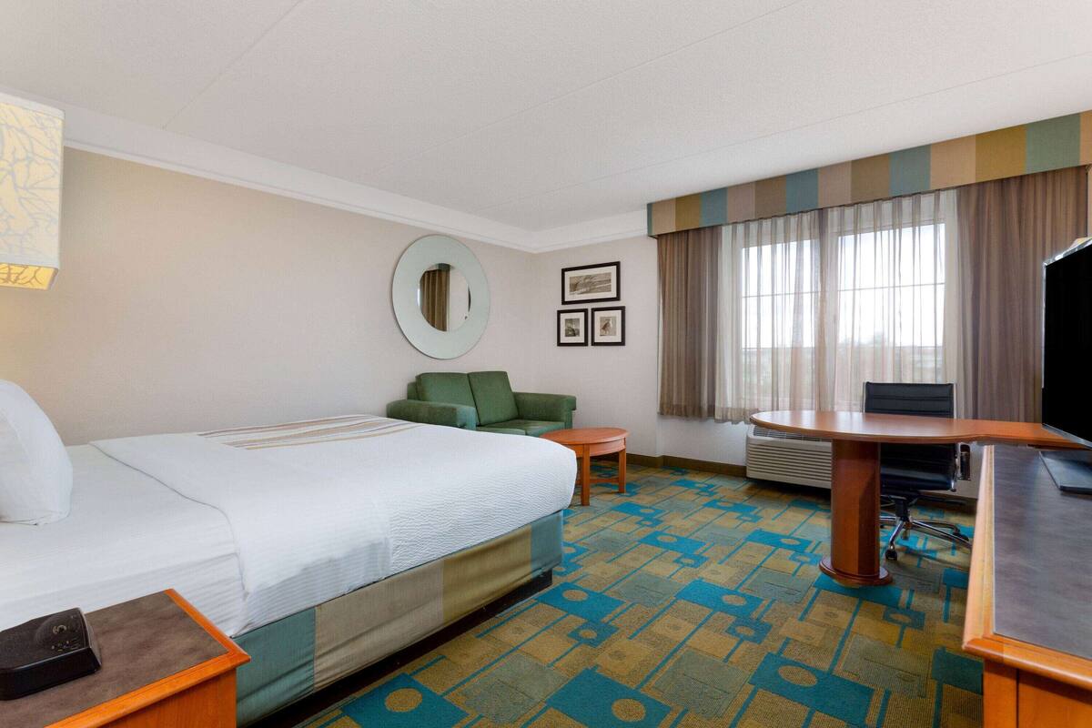 A hotel room with a bed, a green armchair, a small table, a desk with a chair, a TV, and a large window with curtains.
