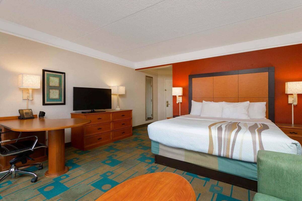 A hotel room with a bed, a desk with a chair, a television, a dresser, and a carpet with a geometric pattern.