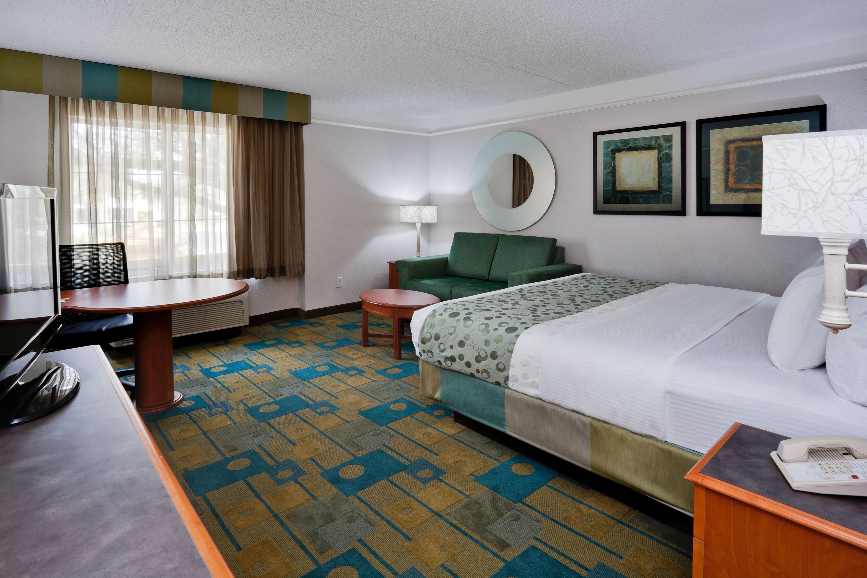 A hotel room with a bed, a desk, a chair, a round mirror, and two framed pictures on the wall.