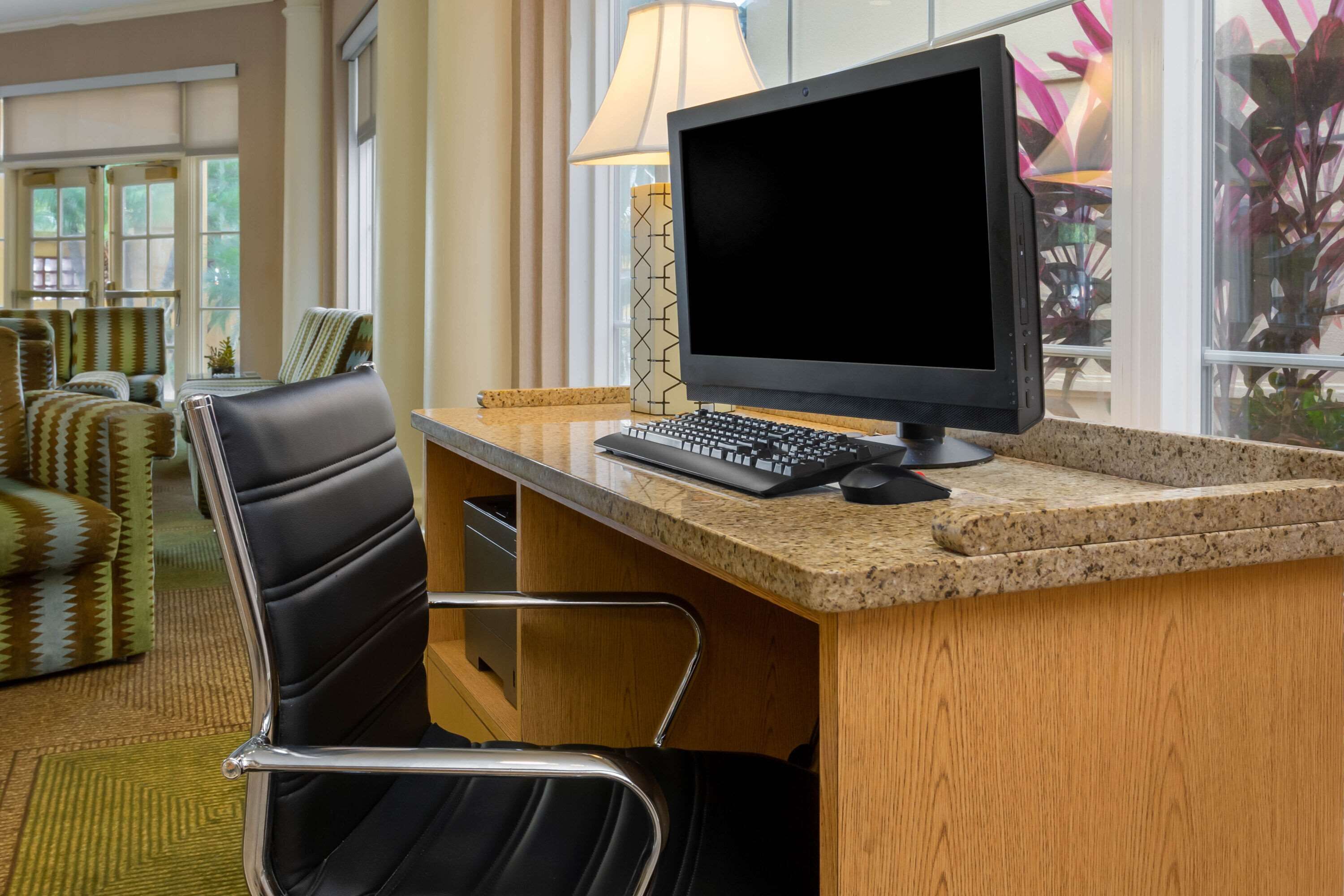 A hotel room with a wooden desk, a black chair, a computer, and a lamp.