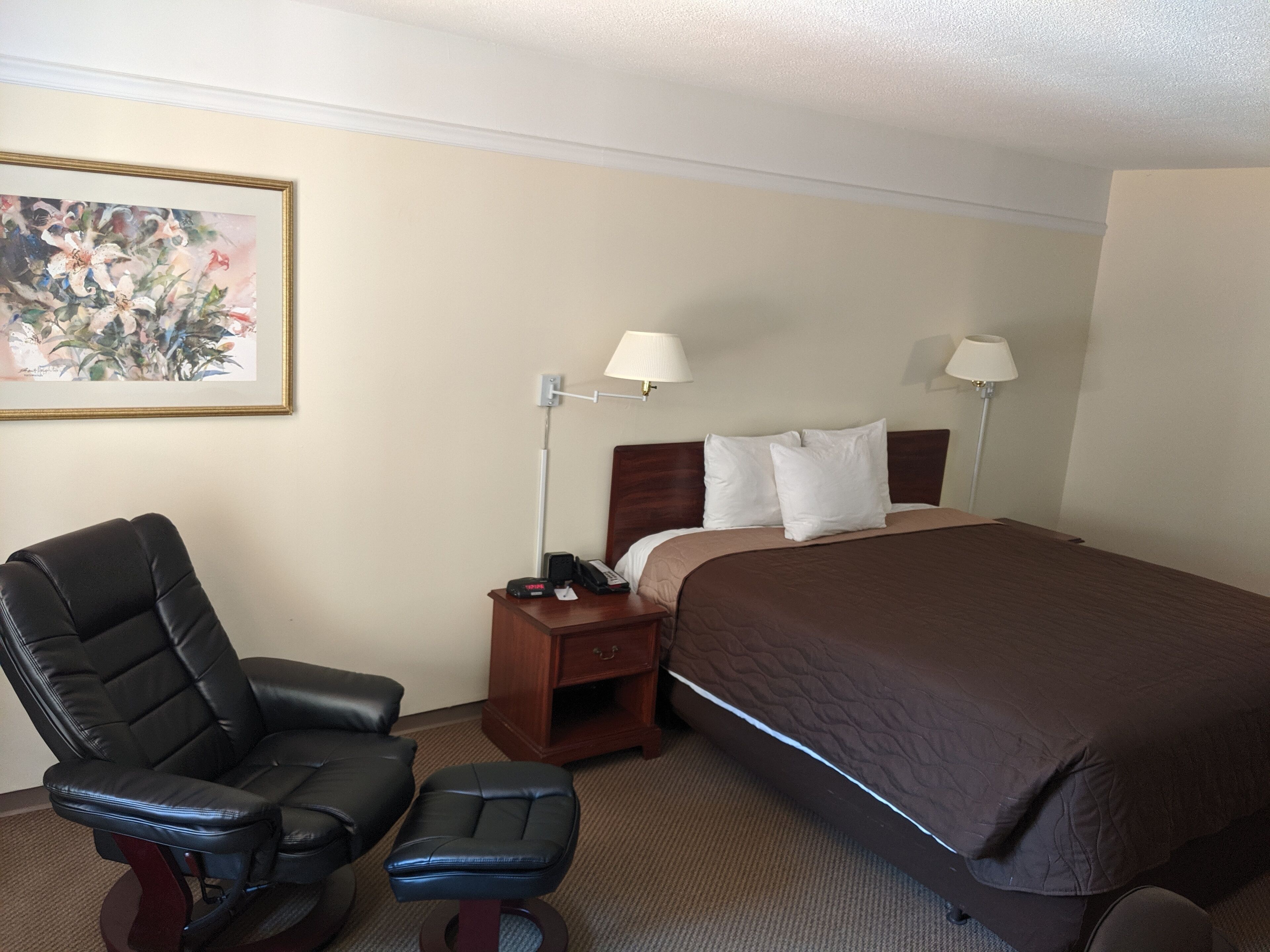 Una habitación de hotel con una cama, una silla, una mesita de noche, una lámpara y un cuadro en la pared.