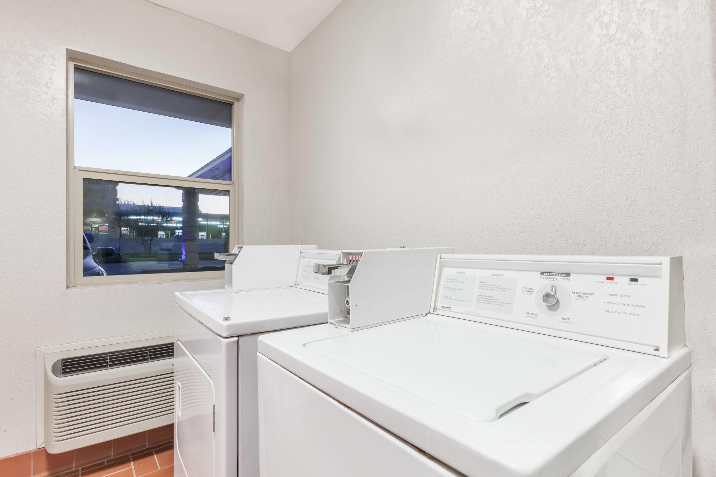 A laundry room with two white washing machines, a vent, and a window with a view of a building outside.