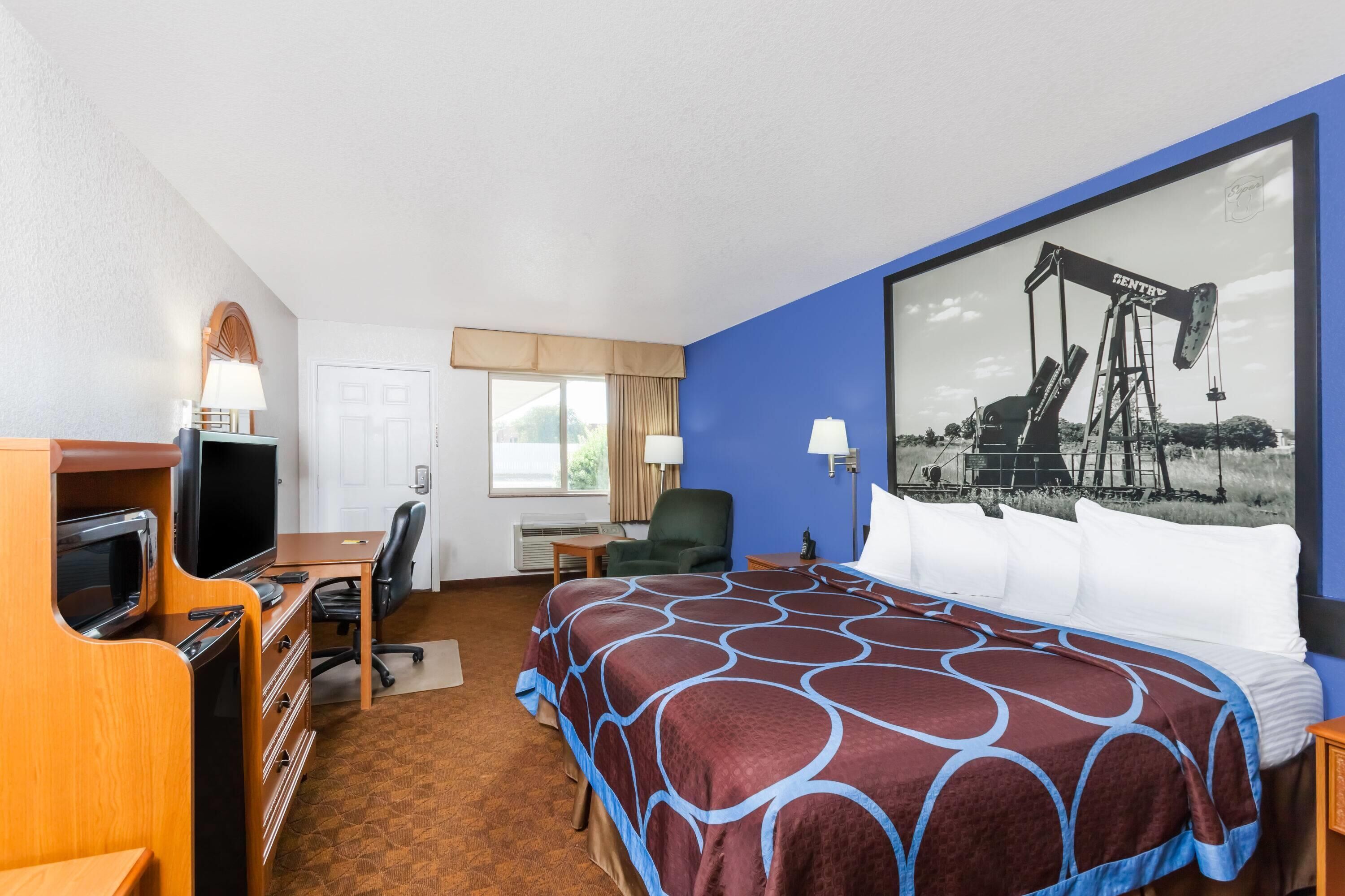 A hotel room with a bed, desk, television, and a large painting of an oil rig.