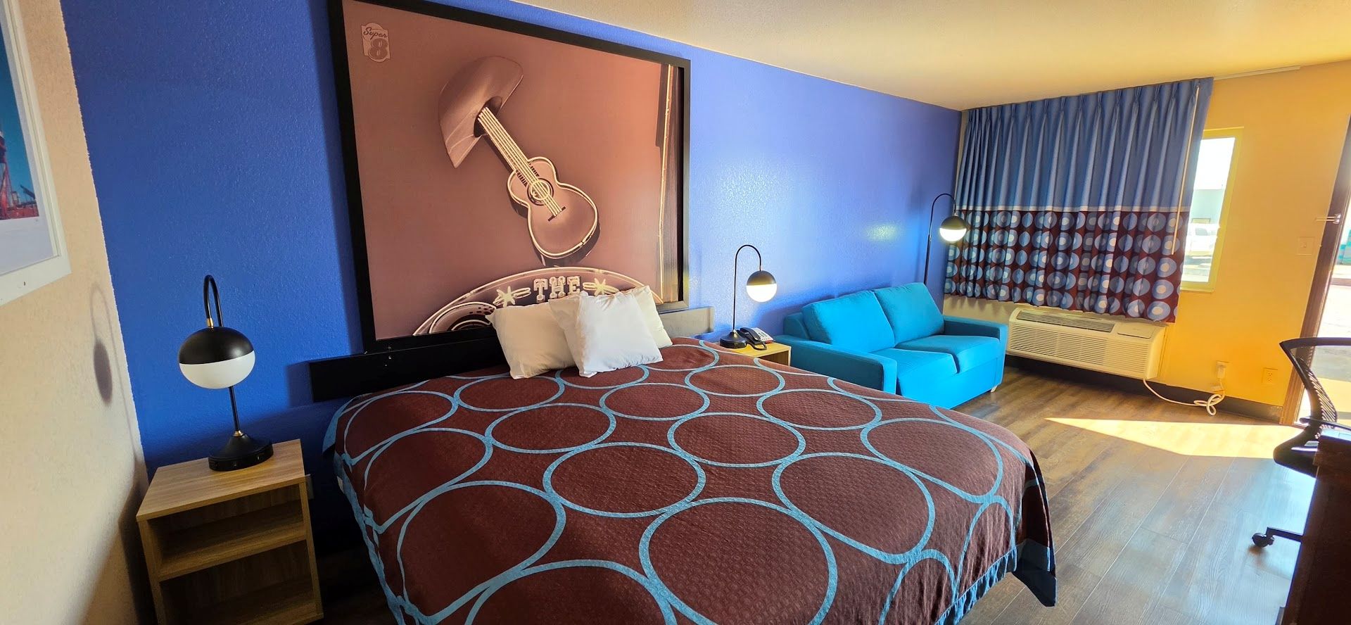 A hotel room with a bed, a blue sofa, a guitar decoration, a lamp, and a window with curtains.