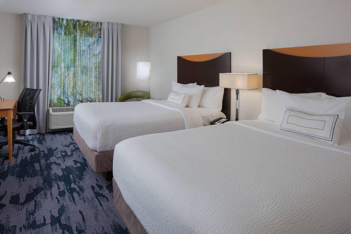 A hotel room with two beds, a desk, a chair, a large window with curtains, and a wall-mounted lamp.