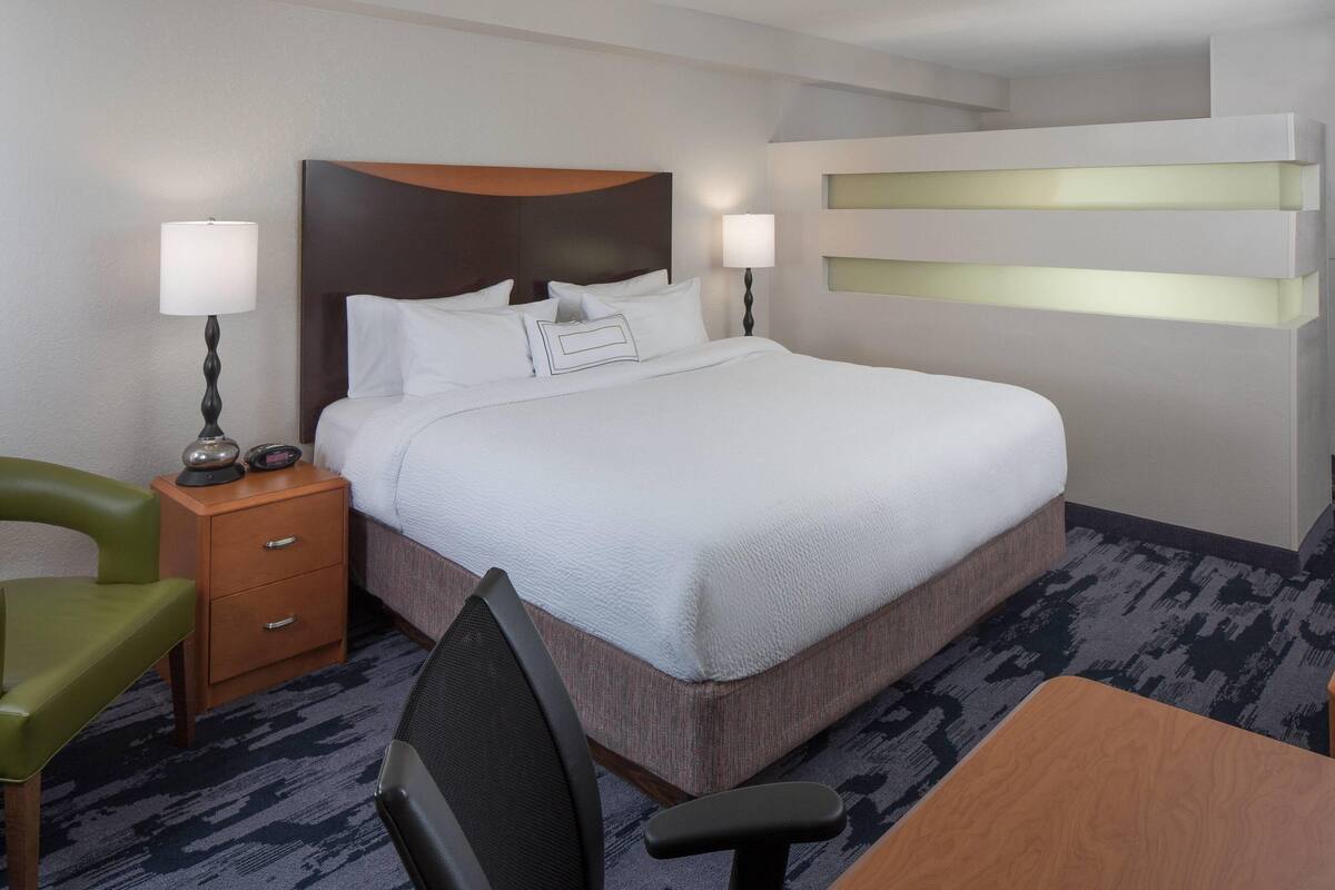 A hotel room with a large bed, two bedside tables with lamps, a green chair, a wooden desk, and a carpeted floor.