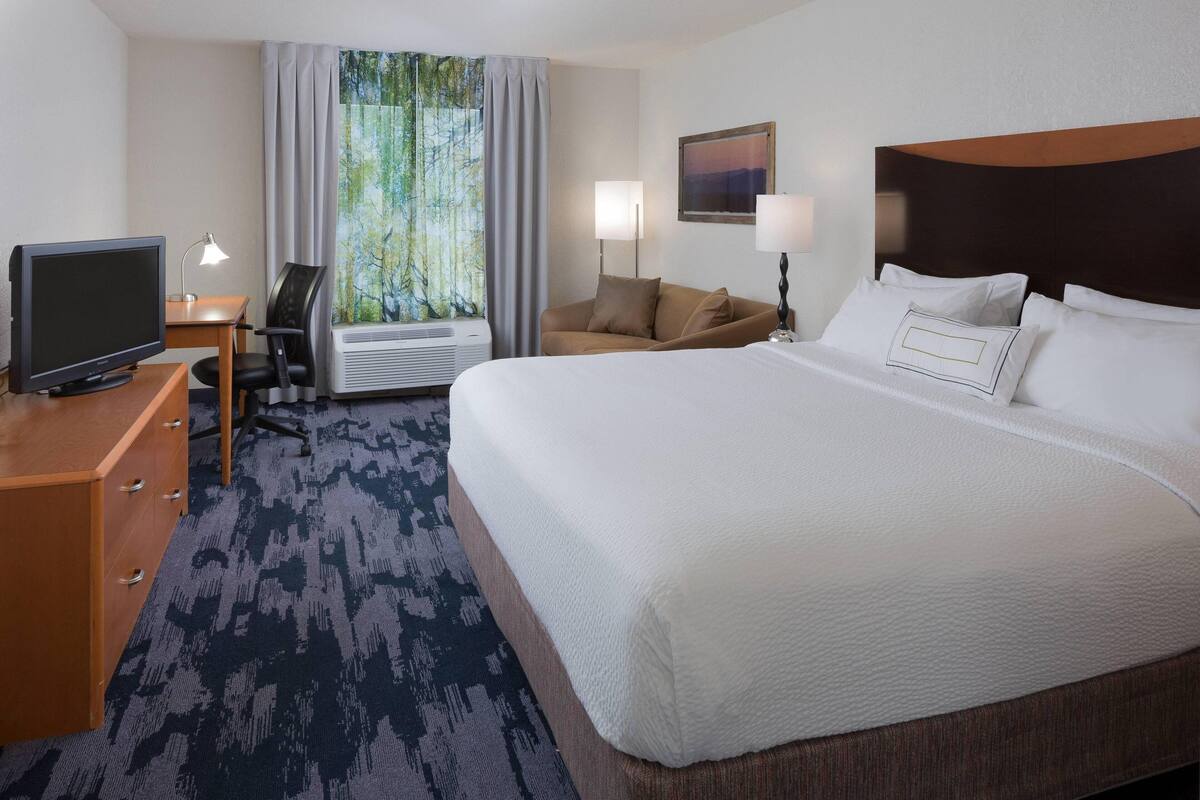 A hotel room with a large bed, a desk with a computer, a chair, a lamp, and a window with curtains.