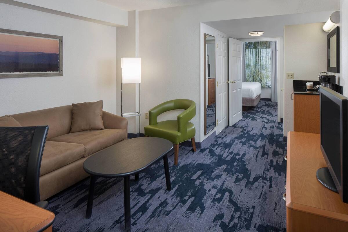 A hotel room with a sofa, a green armchair, a small table, a television, and a desk with a mirror.