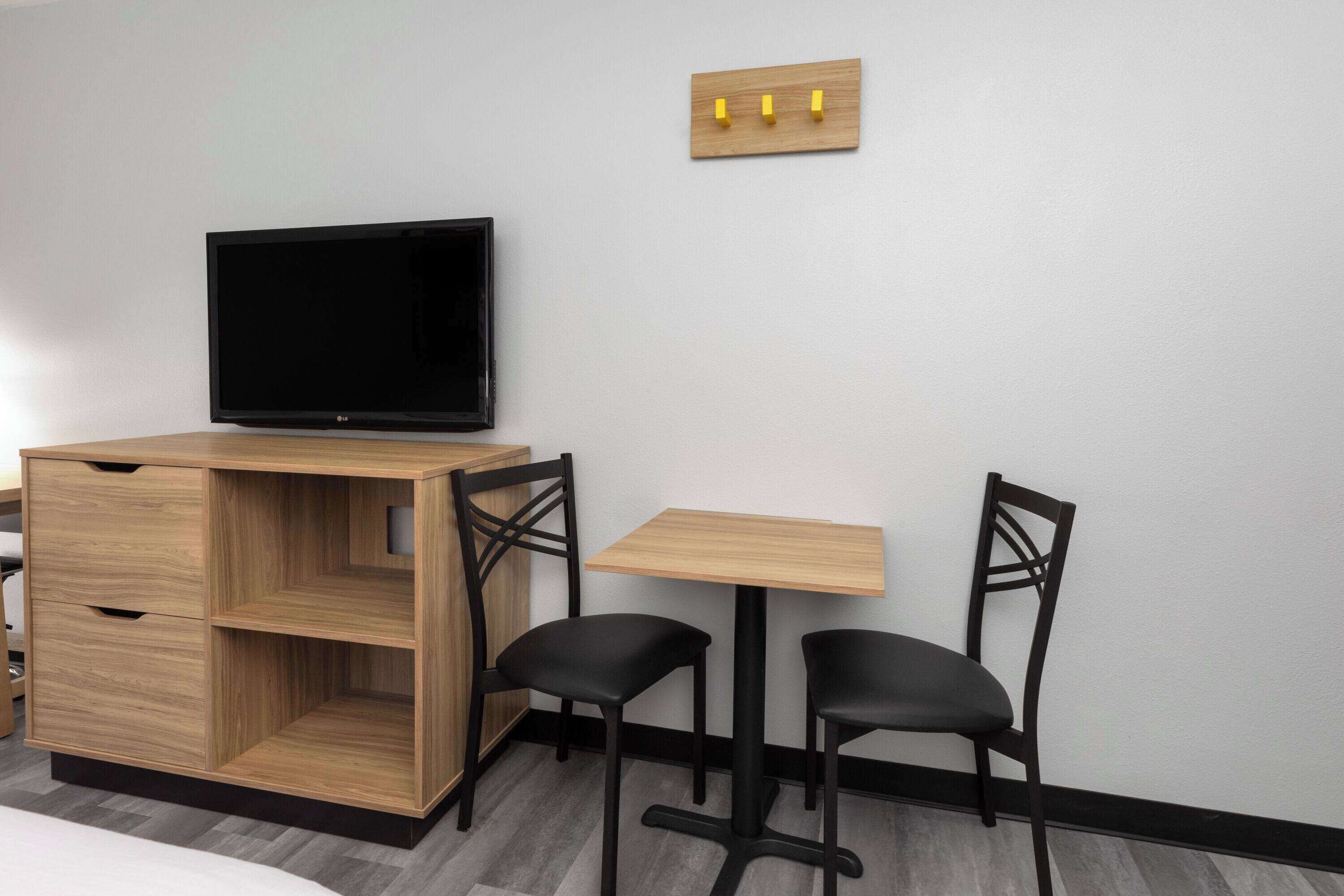Une pièce de taille modeste, avec un téléviseur posé sur un meuble en bois, deux chaises noires, une petite table en bois et un interrupteur fixé au mur.