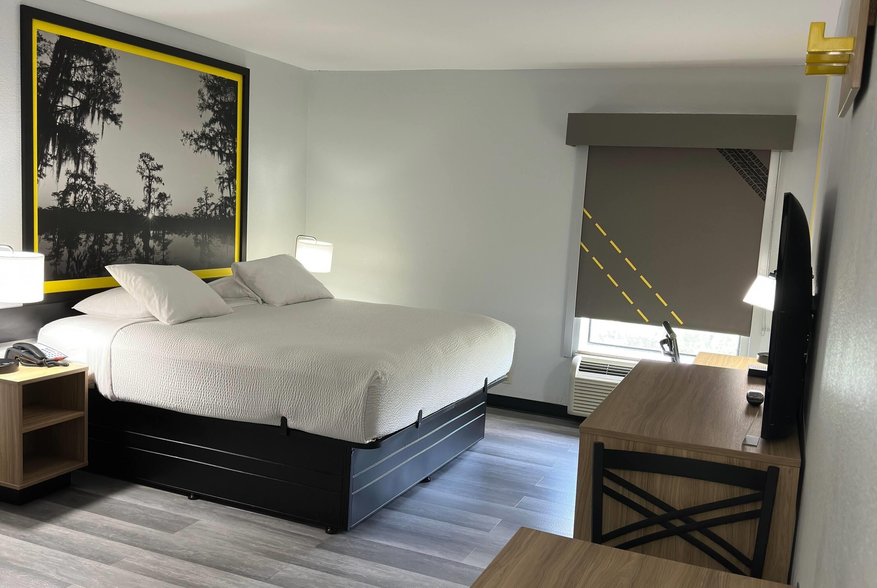 A modern hotel room with a large bed, bedside tables, a desk, and a large window with blinds.