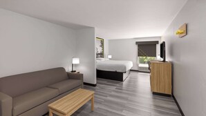 Una habitación de hotel moderna con un sofá, una mesa de madera, una cama, un escritorio con una computadora y un ventanal con vistas a árboles.