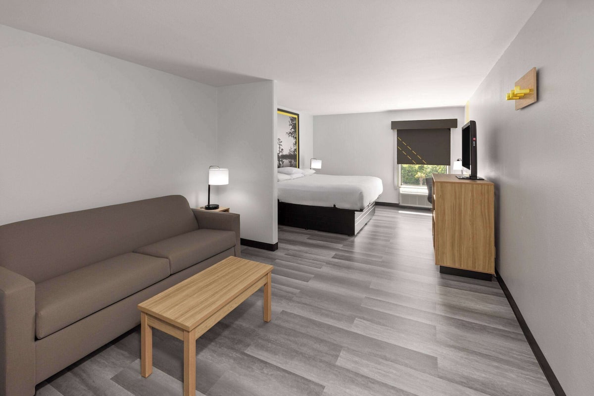 Una habitación de hotel moderna con un sofá, una mesa de madera, una cama, un escritorio con una computadora y un ventanal con vistas a árboles.