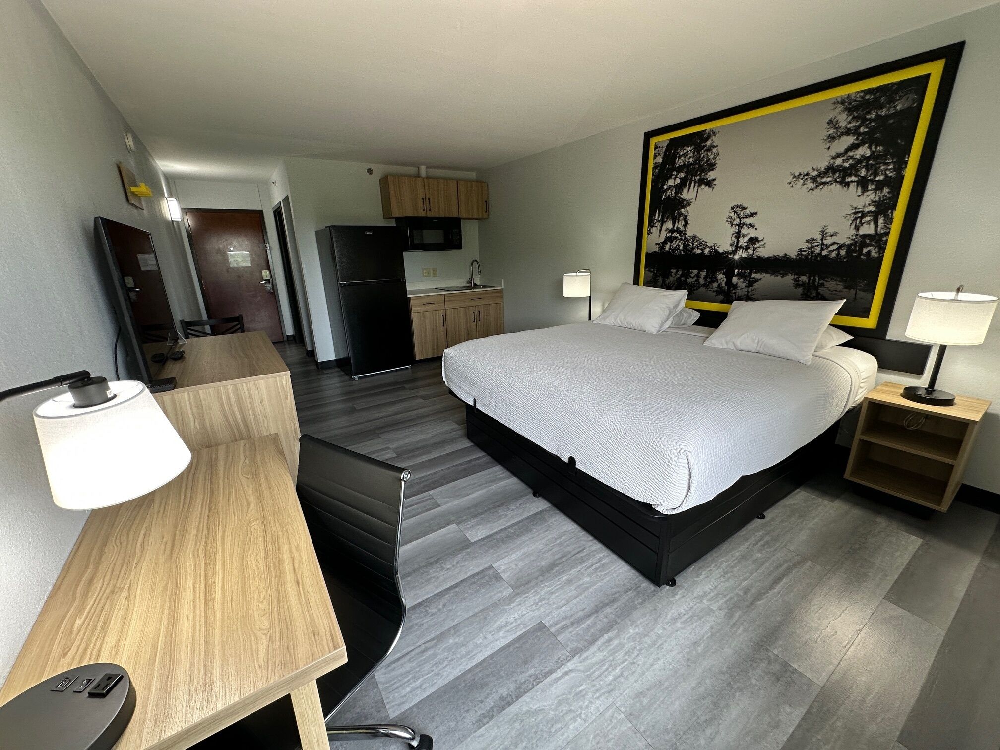 Habitación de hotel con cama, mesitas de noche, escritorio, silla, televisor, refrigerador, microondas y una zona de cocina.
