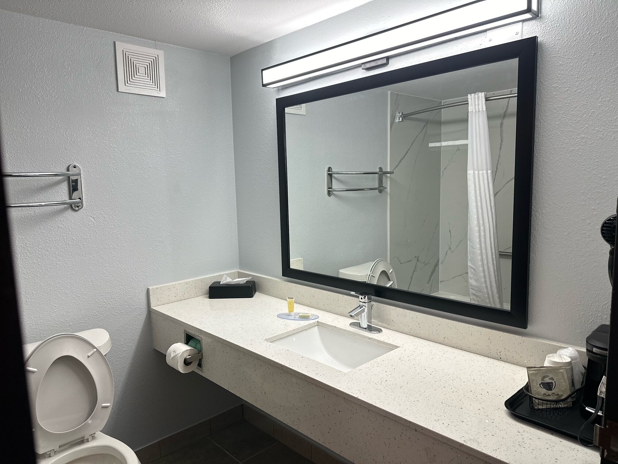 A bathroom with a white toilet, a sink with a mirror above it, a towel rack, and a coffee maker on the counter.