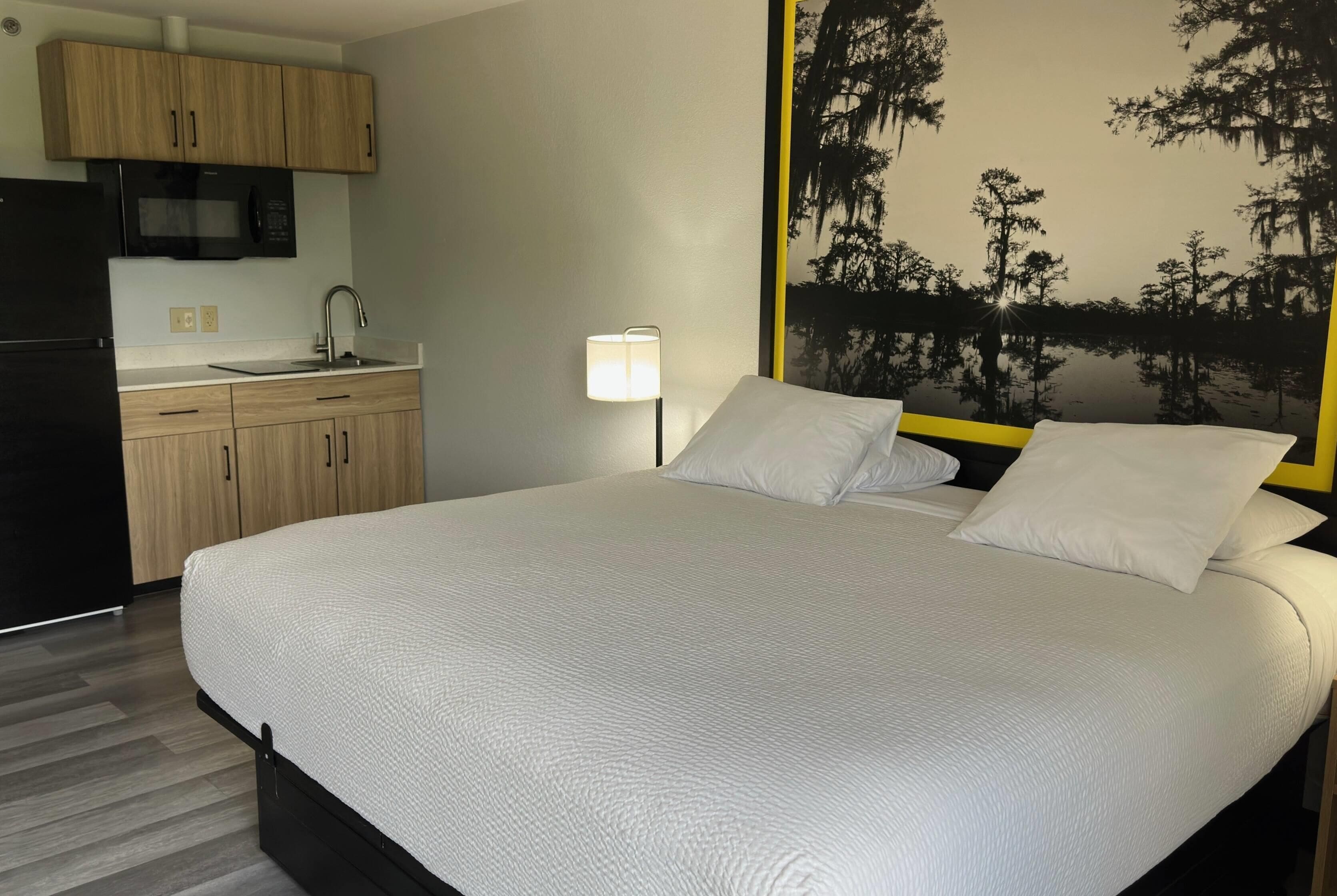 A bedroom with a bed, a nightstand, a lamp, a painting of trees, and a kitchenette with a microwave and refrigerator.