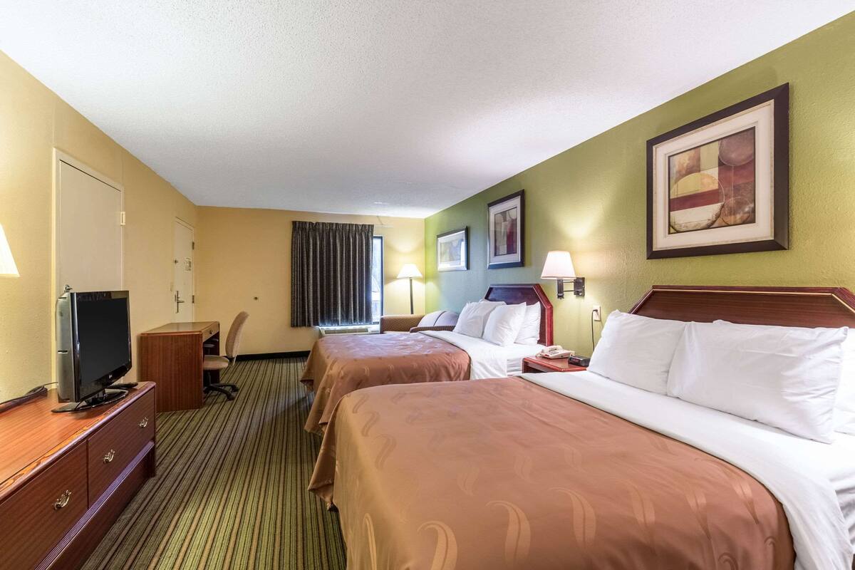 Ein Hotelzimmer mit zwei Betten, einem Schreibtisch mit Fernseher, einem Sessel und einem Fenster mit Vorhängen.