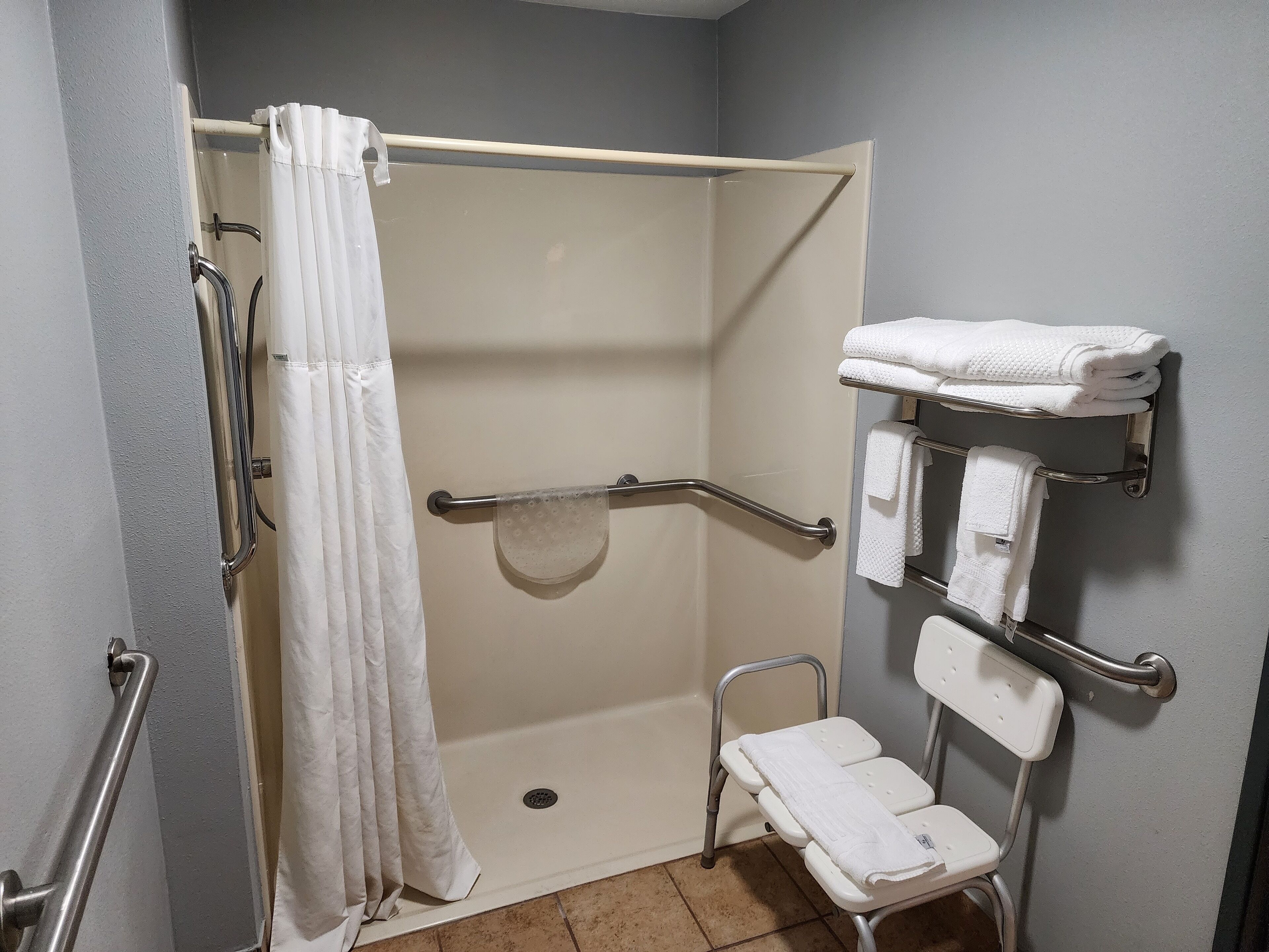 A bathroom with a shower, grab bars, a white towel rack, and a white chair.