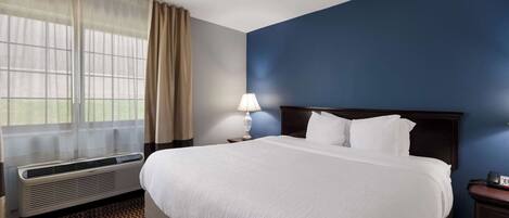 A hotel room with a large bed, a bedside table with a lamp, a window with curtains, and a wall heater.