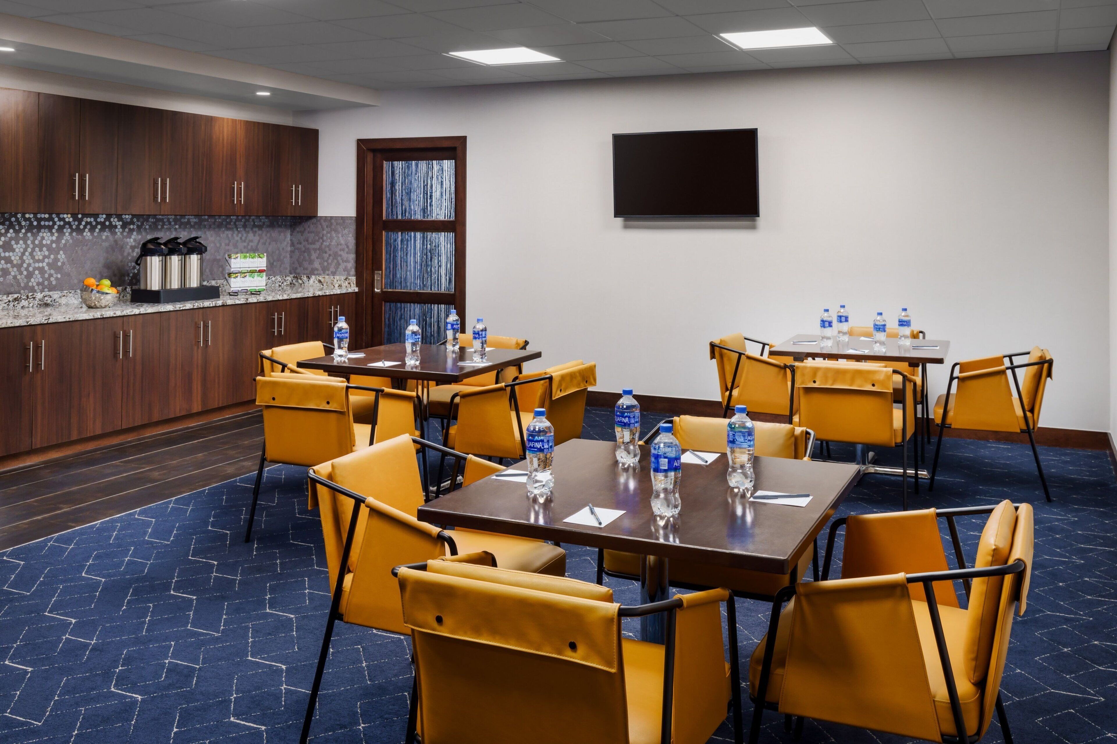 Un salón de conferencias con una gran mesa, sillas amarillas y botellas de agua sobre la mesa. Se observa una zona de cocina con armarios y una encimera.