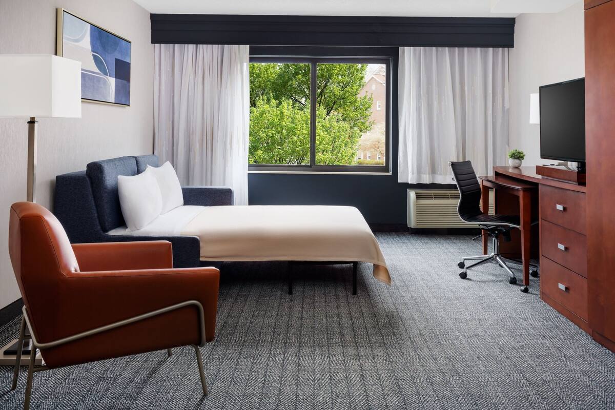 A hotel room with a bed, a desk, a chair, a TV, and a window with curtains.