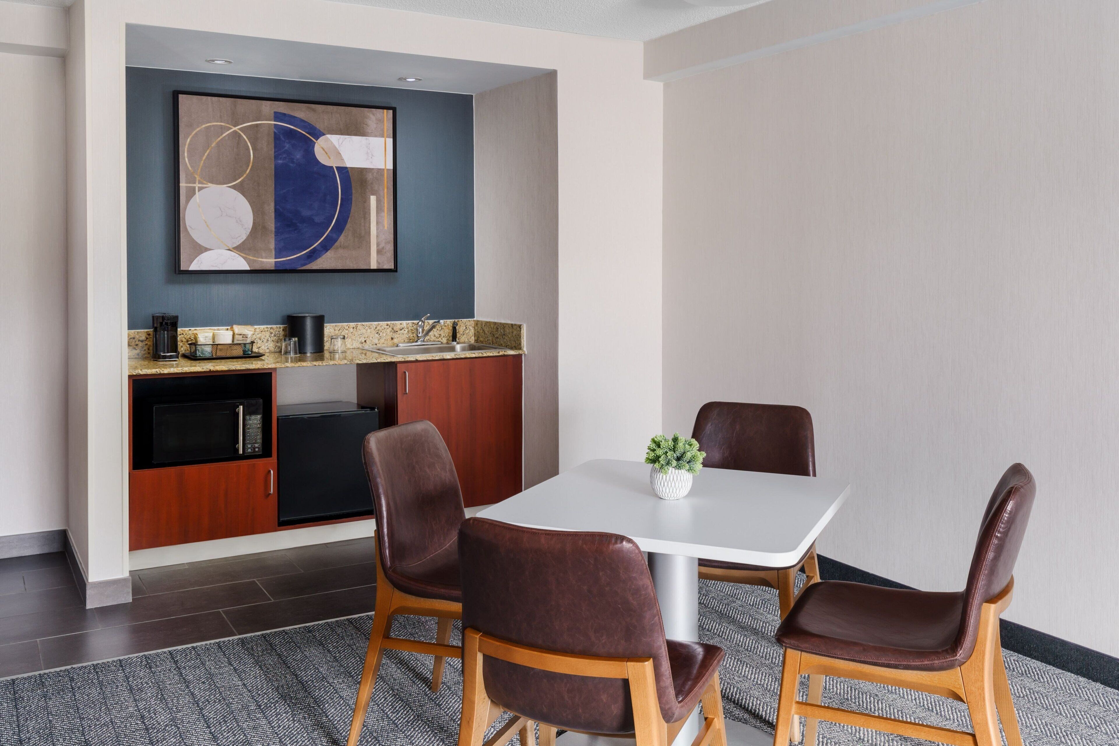A modern hotel room with a dining area featuring a white table, four brown leather chairs, a small potted plant, and a microwave in the kitchenette.