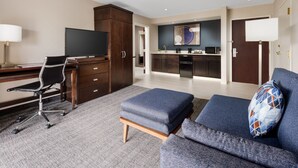 Una habitación de hotel moderna con escritorio, silla de oficina, sofá, reposapiés y una kitchenette al fondo.