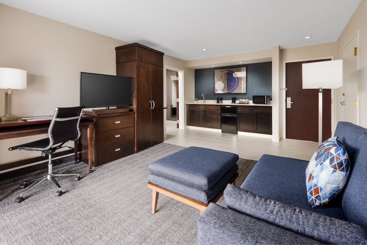 Une chambre d’hôtel moderne équipée d’un bureau, d’un fauteuil de bureau, d’un canapé, d’un pouf et d’une kitchenette à l’arrière-plan.