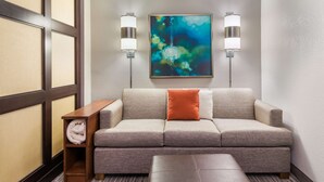 A hotel room with a sofa, a wooden side table with a rolled-up towel, and two wall-mounted lamps.