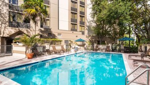 A swimming pool with lounge chairs and umbrellas, surrounded by a building with large windows and a tiled deck.