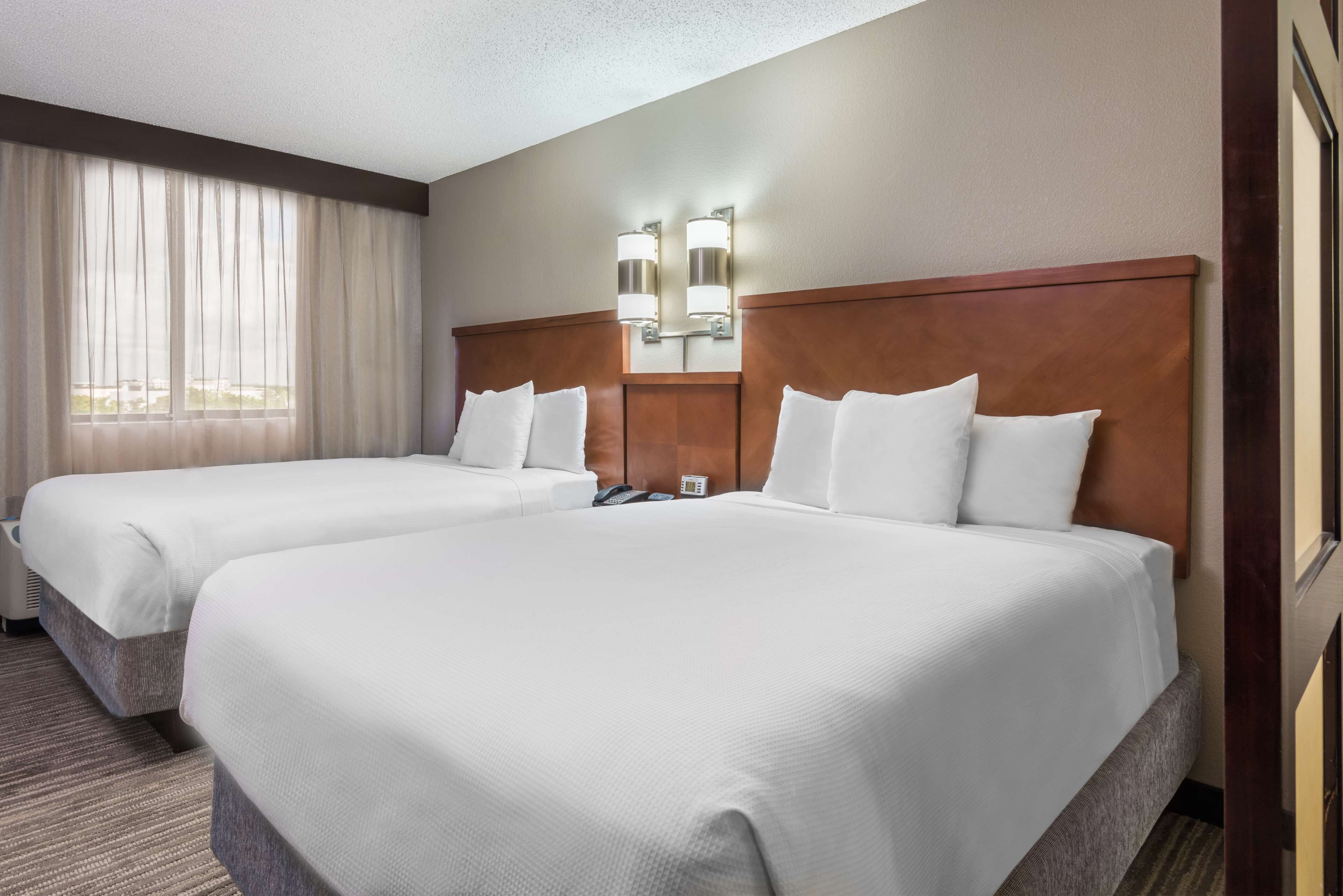 A hotel room with two beds, a large window with sheer curtains, a wooden headboard, and wall-mounted lights.
