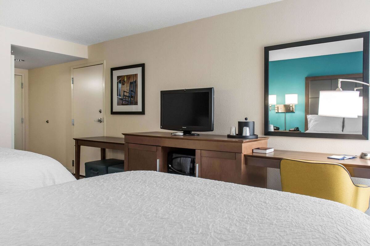 A hotel room with a bed, a desk with a television, a chair, and a large mirror.