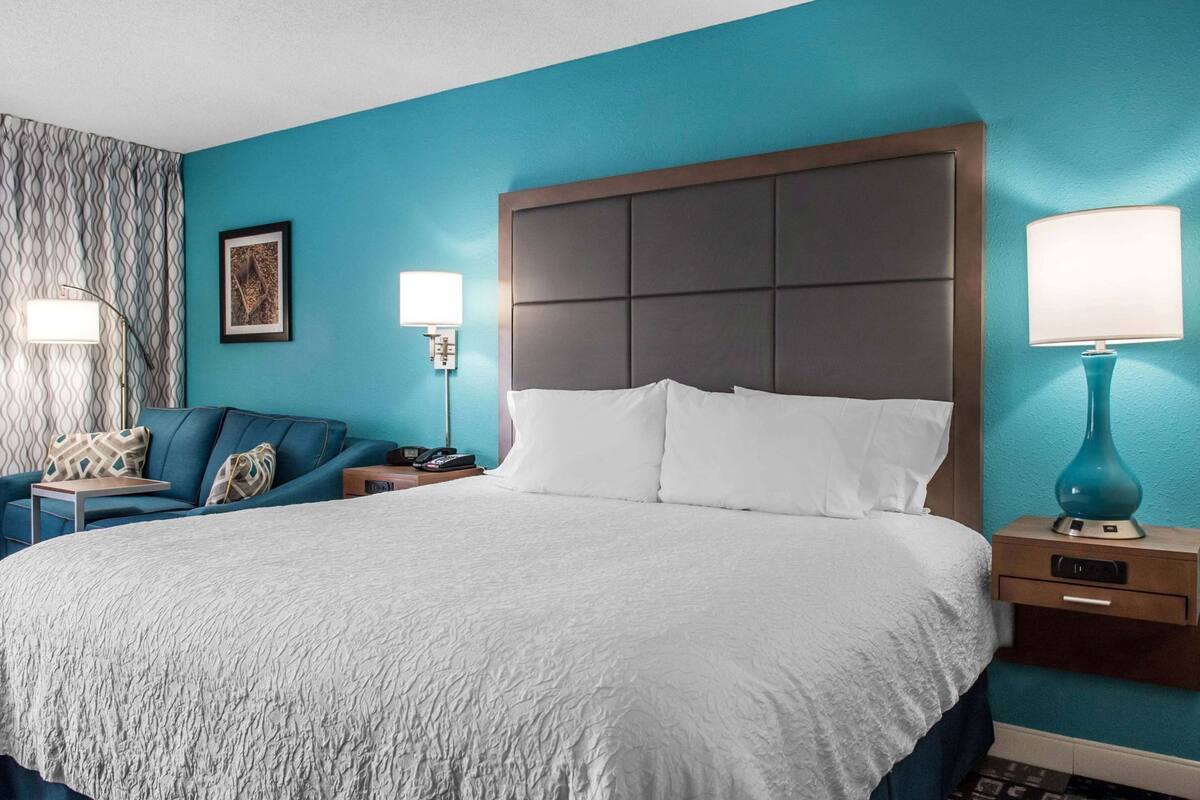 A hotel room with a large bed, a blue sofa, two bedside tables with lamps, and a patterned curtain.