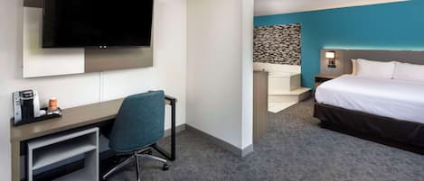 A hotel room with a flat-screen TV mounted on the wall, a desk with a chair, a bed with white bedding, and a bathroom with a blue accent wall.