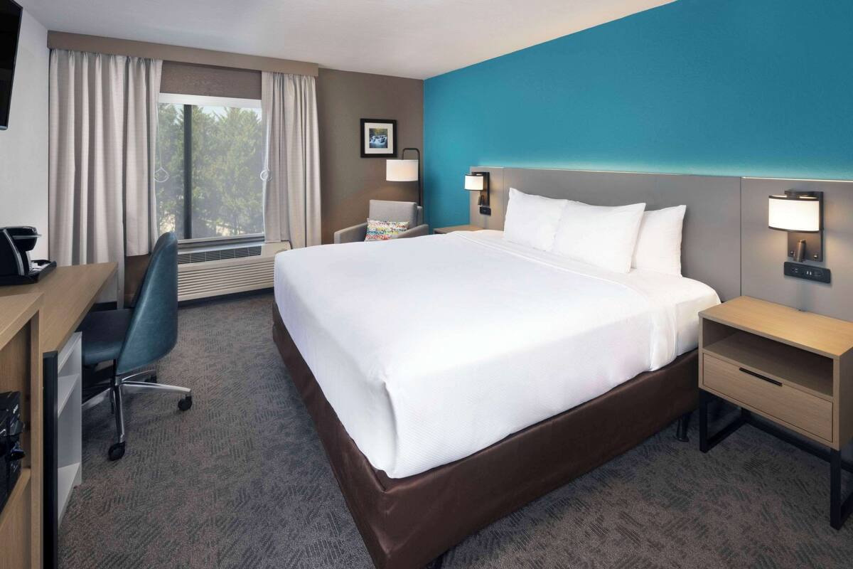 A hotel room with a large bed, a desk, a chair, a desk lamp, a nightstand, and a window with curtains.