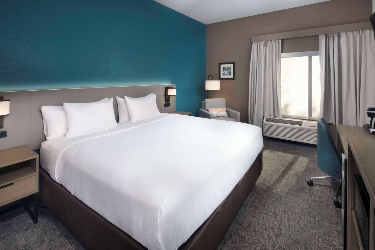 A hotel room with a large bed, a desk, a chair, a window with curtains, and a wall-mounted air conditioner.