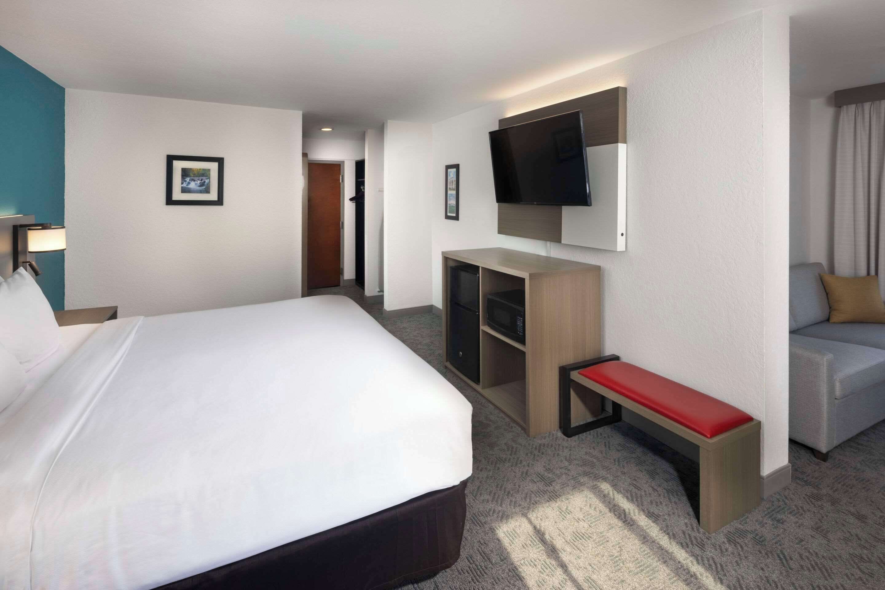 A hotel room with a large bed, a TV mounted on the wall, a seating area with a bench, and a small desk.