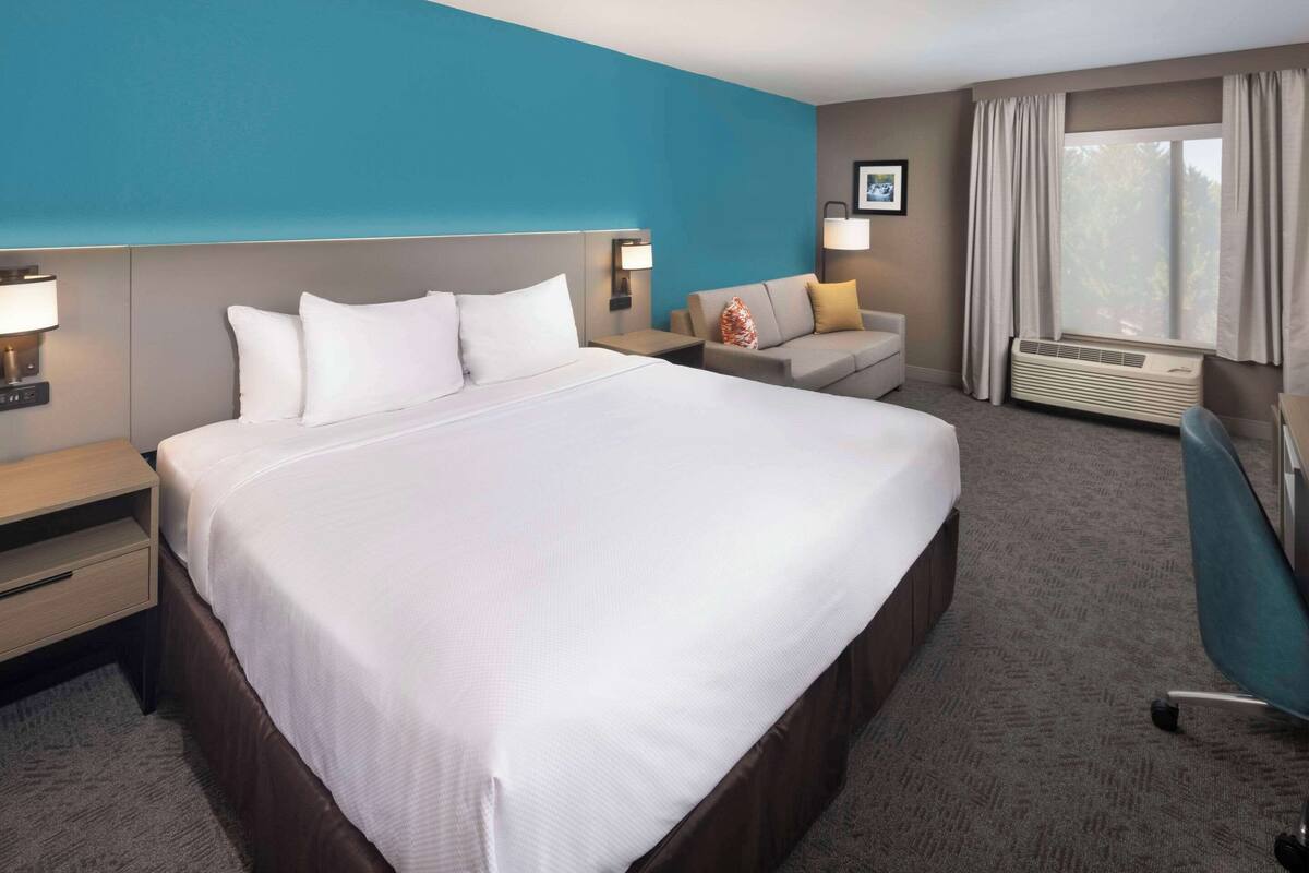 A hotel room with a large bed, a desk, a chair, a sofa, a window with curtains, and a wall-mounted air conditioner.