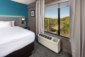 A hotel room with a large bed, a nightstand, a lamp, a window with a view of greenery, and a wall-mounted air conditioner.