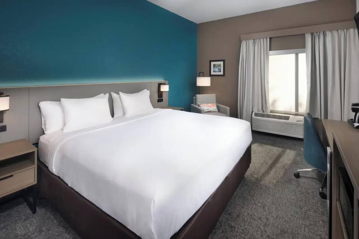 A hotel room with a large bed, a desk, a chair, an air conditioning unit, and a window with curtains.